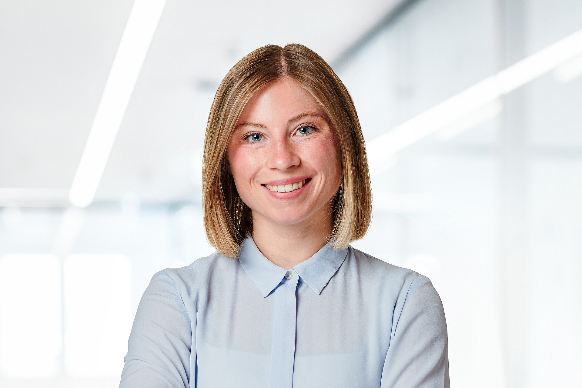 [Translate to Deutsch:] A smiling individual stands in a modern, brightly lit office setting.