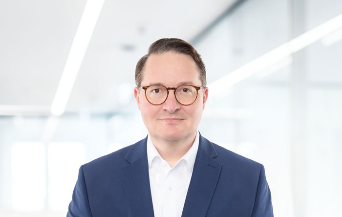 Porträt eines Mannes mit Brille und einem blauen Anzug vor einem hellen Bürohintergrund.