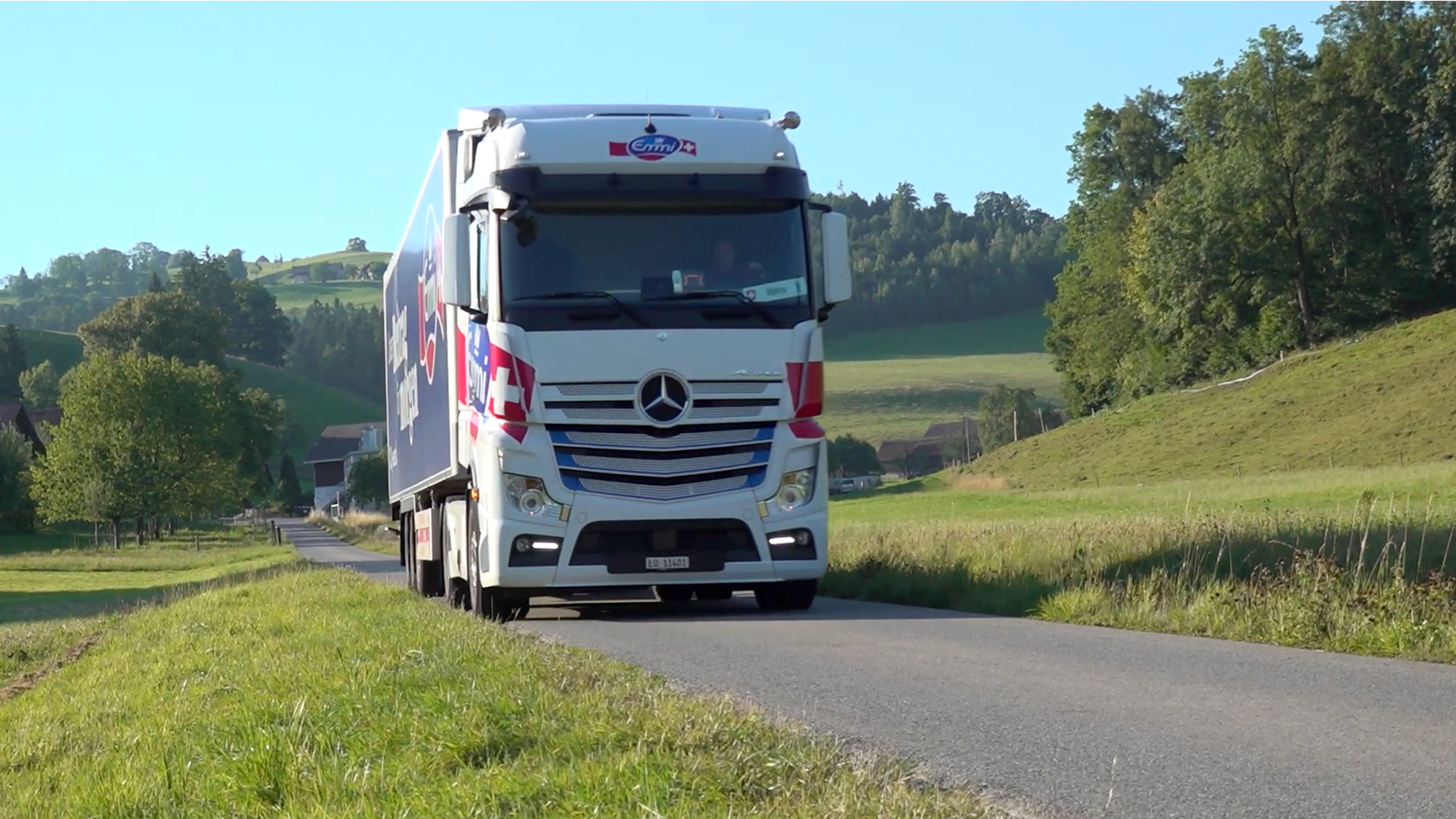 LKW fahren auf einer ländlichen Straße mit grünen Hügeln im Hintergrund.