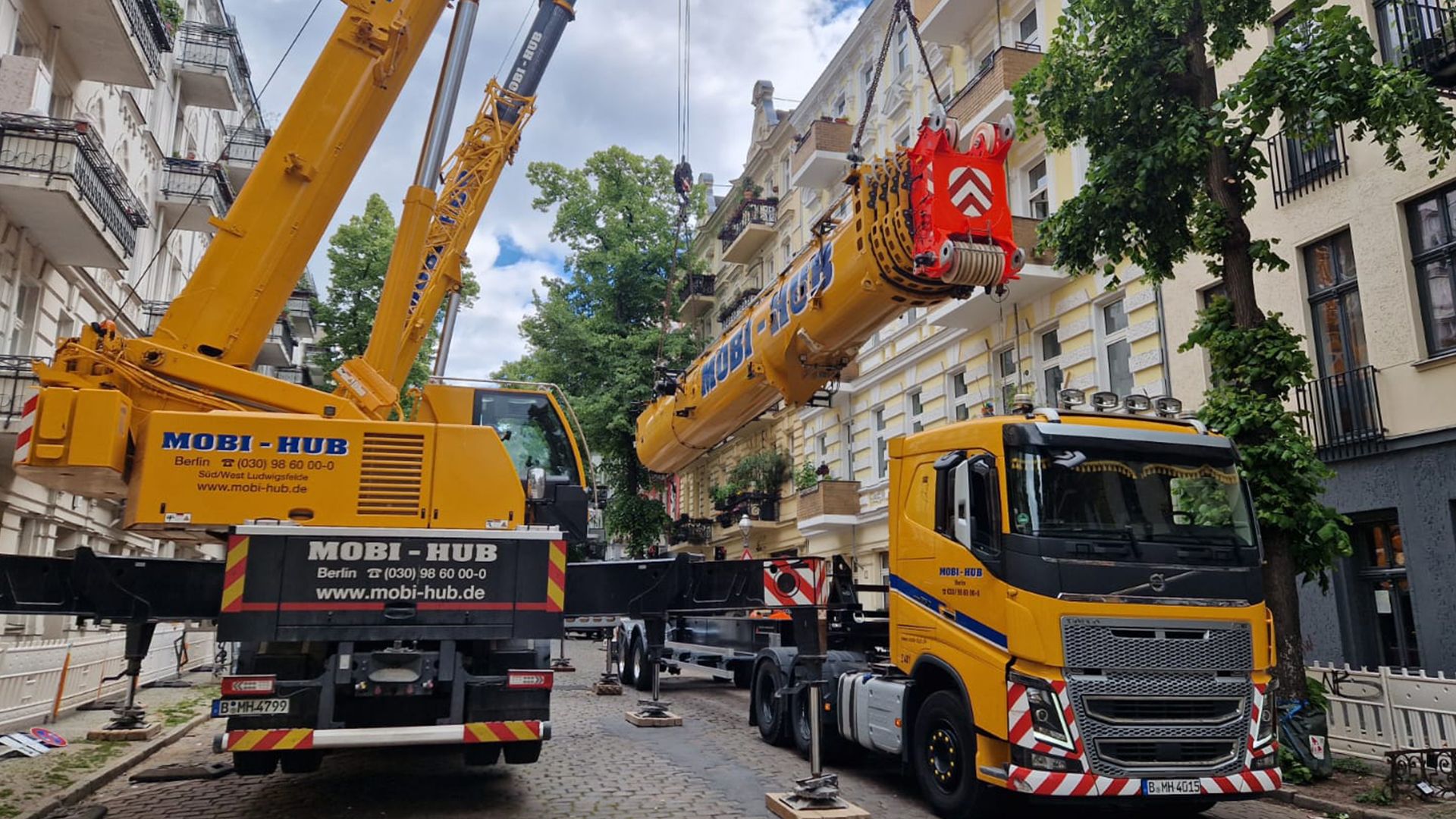Cranes lifting equipment onto a truck in a residential street, with "MOBI-HUB" branding visible.