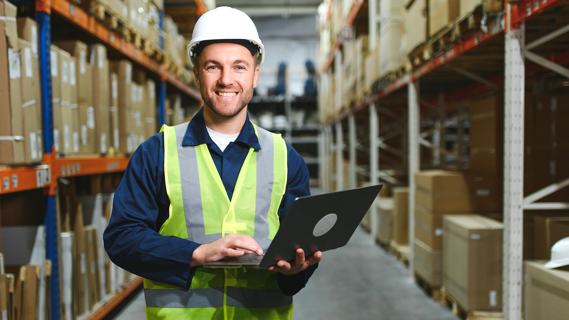 Worker in a hard hat and safety vest using a laptop in a warehouse aisle.