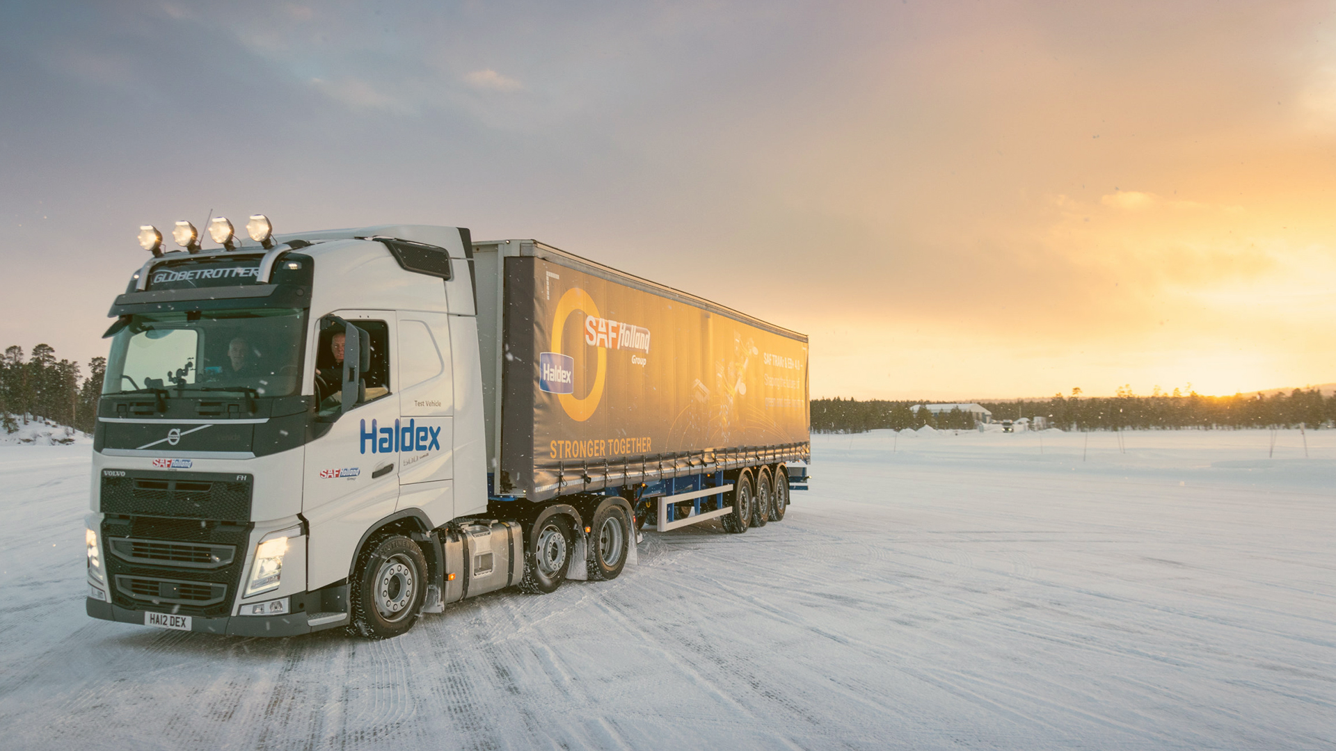 Truck with SAF-HOLLAND branding drives on snowy terrain at sunset.