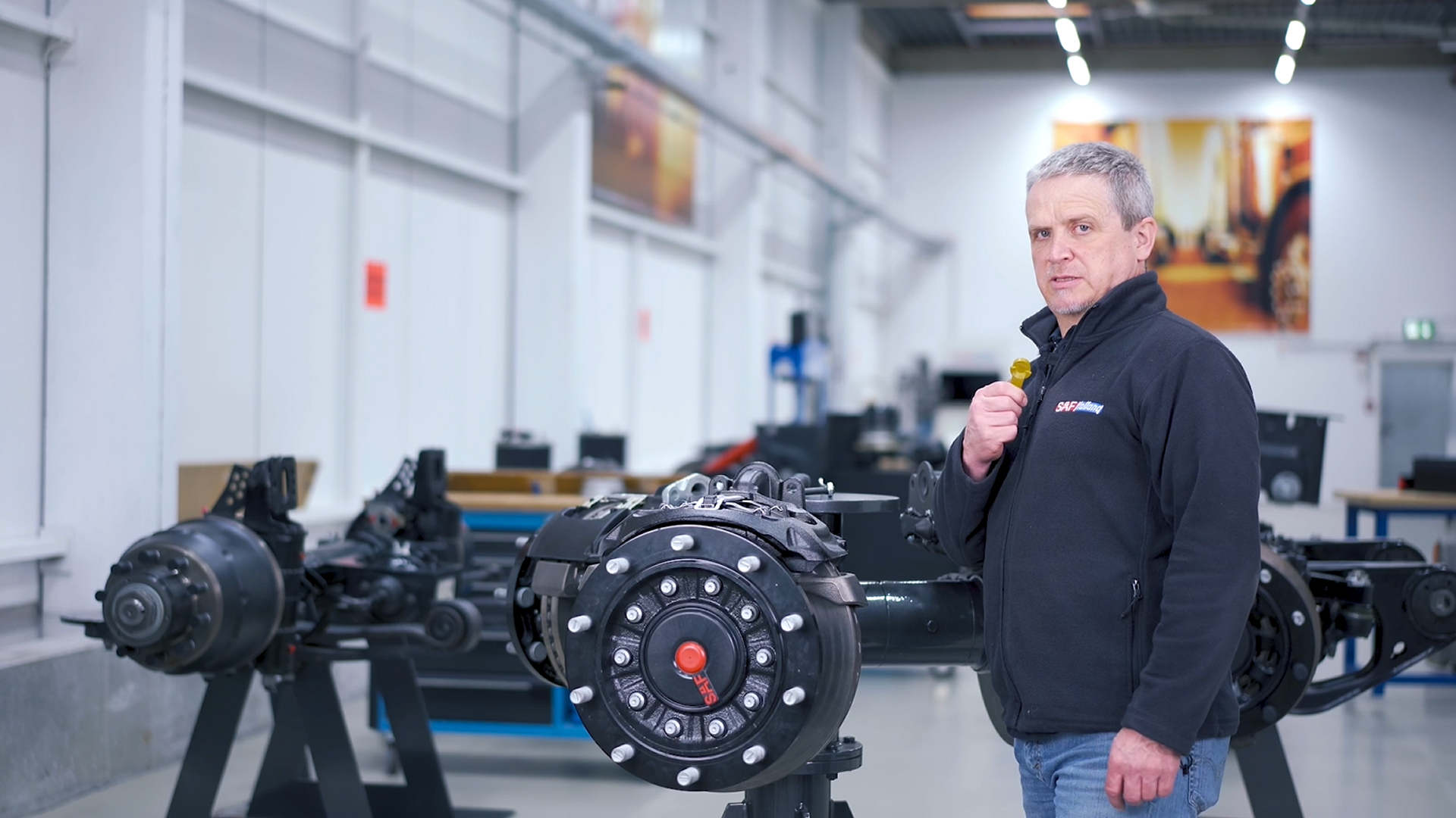 A trainer in a SAF-HOLLAND jacket stands by an axle system in an industrial facility.