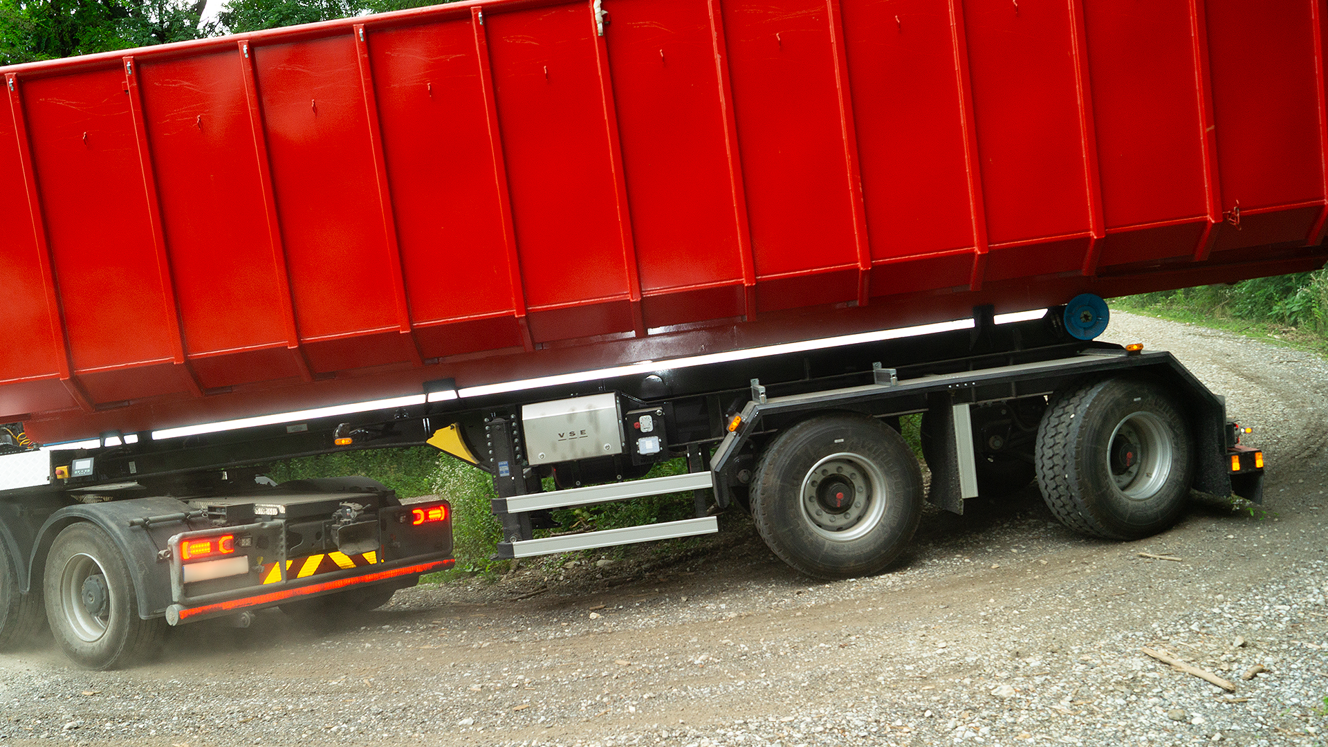 Truck trailer tipping red container on gravel road, showcasing axles and suspension.
